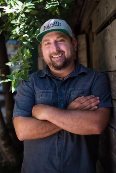 Man in a cap and blue shirt, smiling outdoors with folded arms.